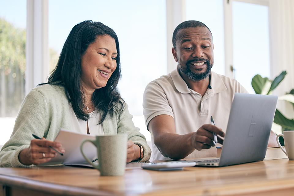 Add More to Your Savings This Year Smiling woman and man sitting at table looking at laptop.