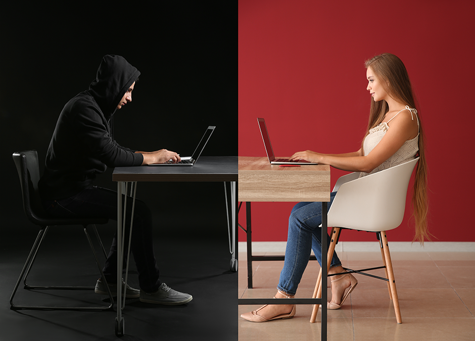 Ominous man sitting at table on computer facing smiling woman sitting at table on a computer.