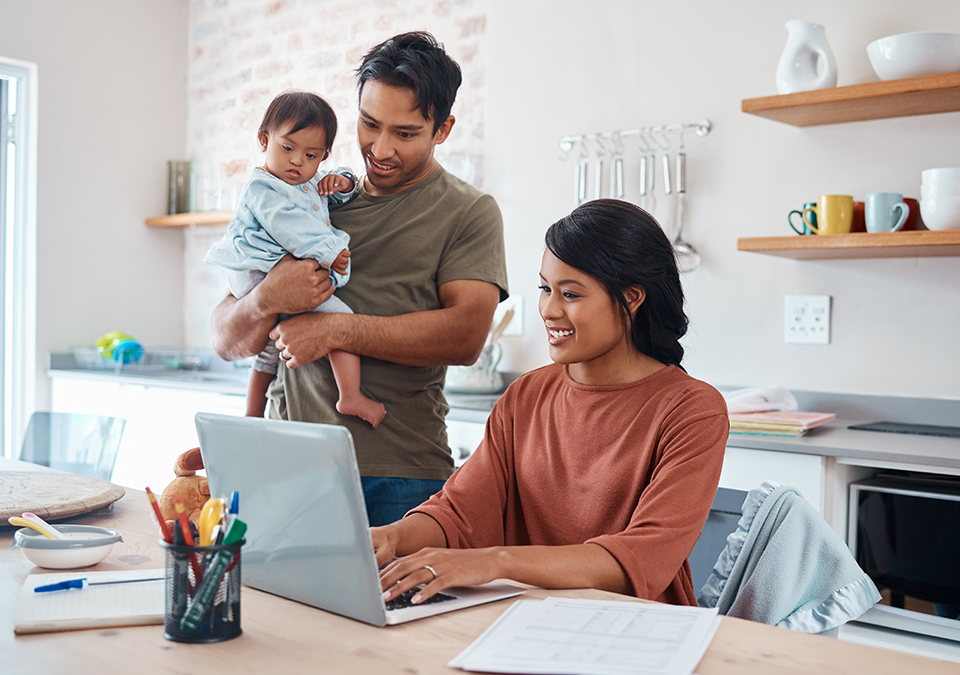 Man holding baby while standing next to woman sitting at table working on laptop