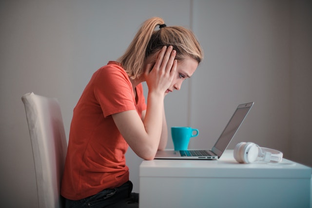 Distressed woman sitting at desk looking at laptop with head in hands.
