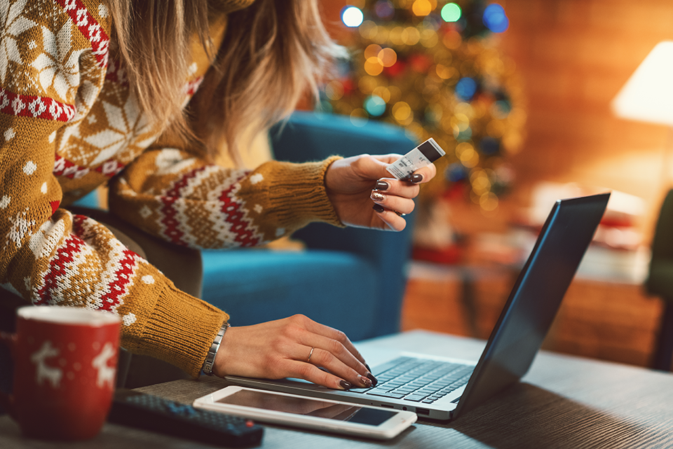 Woman sitting on couch in holiday sweater shopping on laptop.