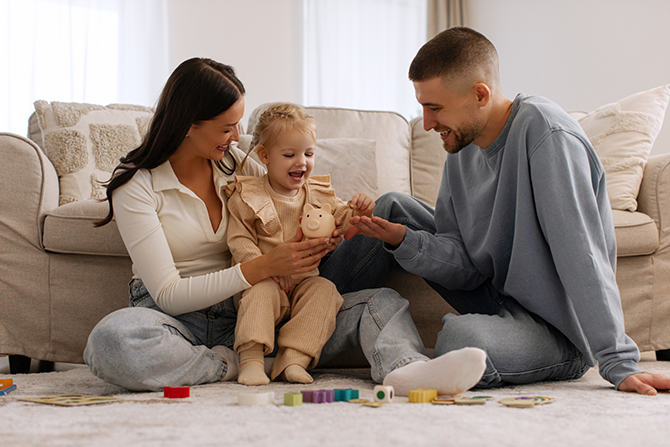 parents sitting on floor with toddler girl playing with piggybank