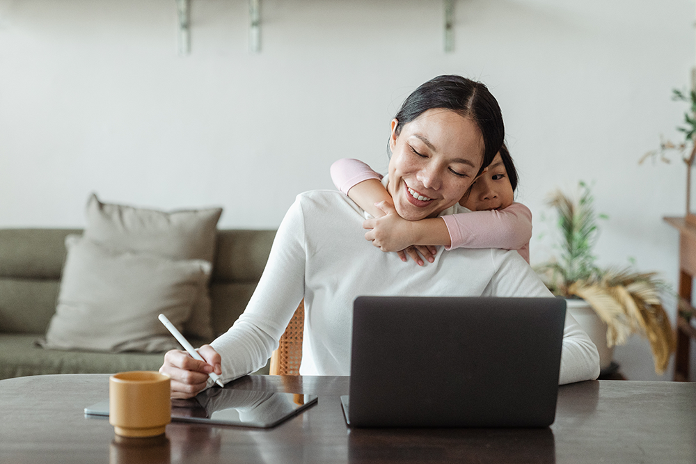 Smiling woman sitting at table working on laptop while child hugs her around the neck.