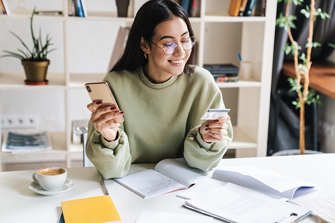 smiling woman sitting at desk looking at credit card while holding phone