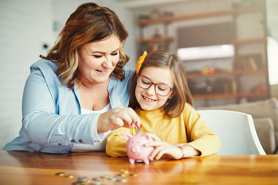 Woman and child sitting at table putting coins in piggy bank.