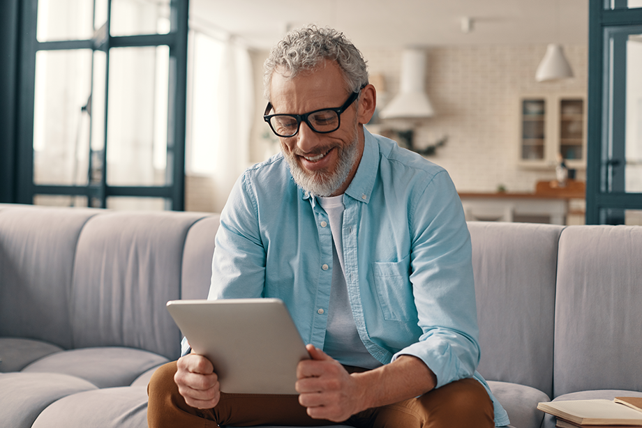 Cheerful senior man in casual clothing using digital tablet while sitting on the sofa at home