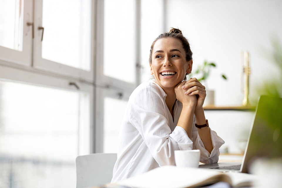 Smiling woman leaning chin on clasped hands sitting at desk.