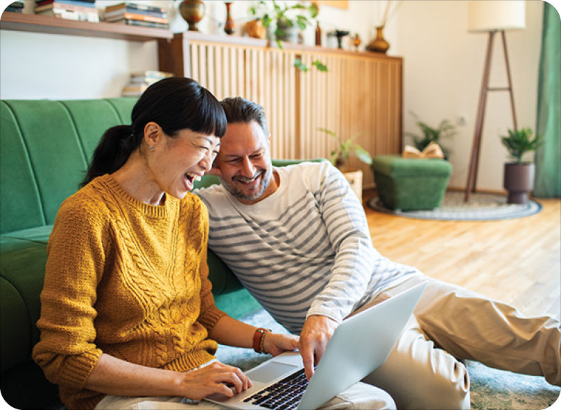 woman and husband sitting on floor in front of green couch laughing and smiling at laptop