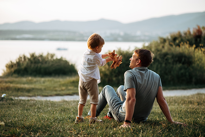 Father sitting in grass while son plays with wooden airplane